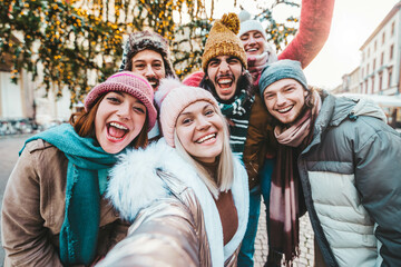 Happy friends group wearing winter clothes taking selfie walking on city street - Cheerful young people hanging outside enjoying winter holidays - Friendship concept with guys and girls laughing loud - obrazy, fototapety, plakaty