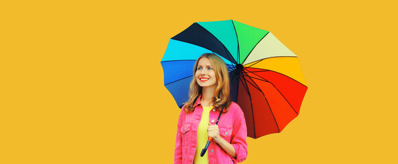 Portrait of beautiful stylish happy smiling young woman holding colorful umbrella wearing pink jacket on bright yellow studio background - obrazy, fototapety, plakaty
