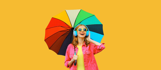 Happy cheerful laughing young woman with colorful umbrella listening to music in headphones wearing pink jacket on yellow studio background