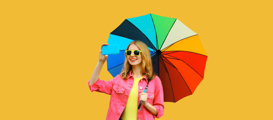 Portrait of stylish happy smiling young woman taking selfie with mobile phone holds colorful umbrella wearing pink jacket on yellow studio background