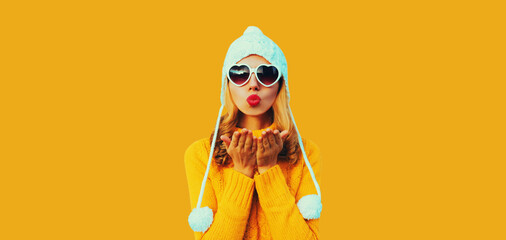 Winter portrait of beautiful woman blowing her red lips sending sweet air kiss wearing heart shaped sunglasses, yellow knitted sweater and white hat with pom pom on studio background - obrazy, fototapety, plakaty