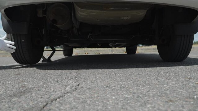 Unrecognizable man in gloves on jacked up car rotates wheel and finds cause of puncture. Wide shot of underbody of broken down car with screw in tire tread on asphalt road on sunny day.