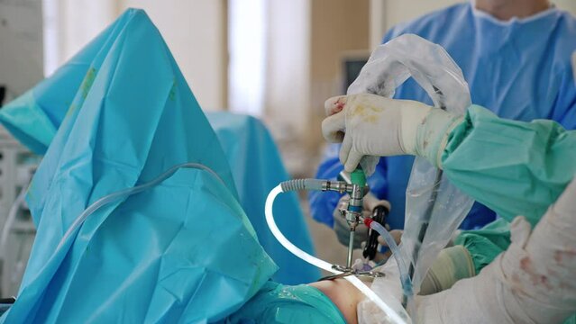 Hands of surgeon in latex gloves stained with blood hold the modern surgical device. Surgeon at backdrop uses chisel and mallet.