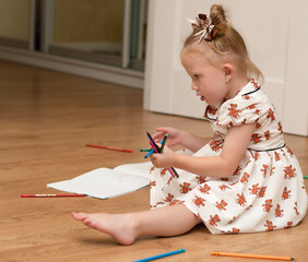 A little beautiful girl in a white dress sits on the floor in a home interior, draws and plays with colorful pencils.