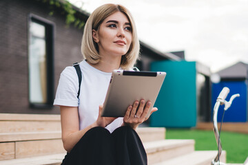 Thoughtful adult modern female sitting on stairs with bicycle browsing tablet in street