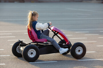 Little girl is riding on pedal karting. Child on a toy pedal car rides on a bike path outdoor,...