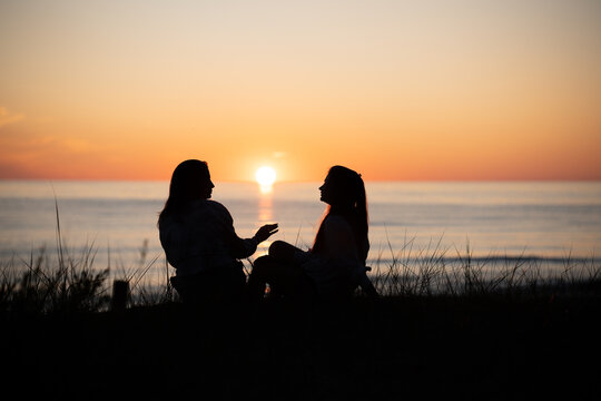Silhouette of two female friends enjoying a conversation while sitting and watching the sunset on a beach. Back view