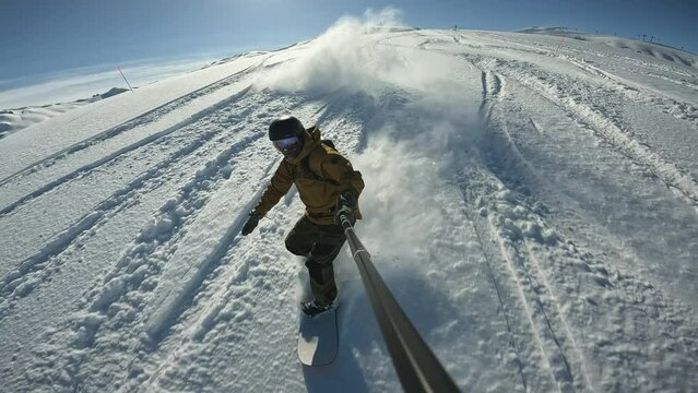 Winter sports activities, man snowboards and filming himself with selfie stick. Snowboarder rides through deep snow and turns in fresh powder. Snow splashes into camera and athlete.