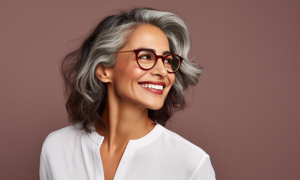 Retrato de una mujer latina madura, con canas, sonriendo, con apariencia saludable y vitalidad, usando una blusa blanca y gafas, posando en un estudio fotogr&aacute;fico con fondo de color