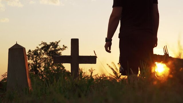 unrecognizable man in black at sunset in cemetery approaches cross and holds on to it. Concept of losing friend or family member. Grief and sadness. Religious funeral ritual.
