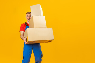 Smiling delivery man or loader holds cartboard boxes on yellow background in studio. Copy space,...