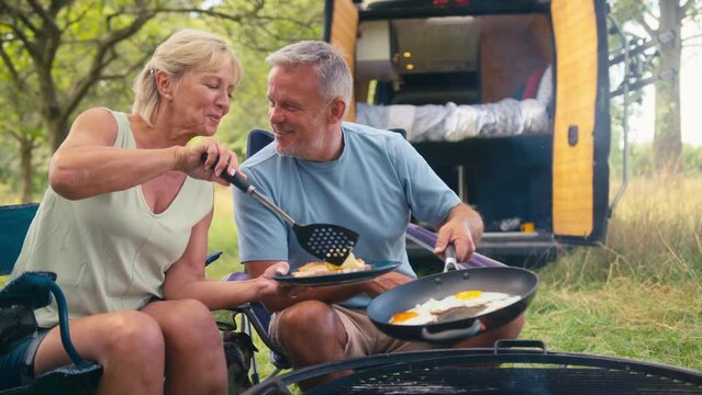 Senior retired couple camping in countryside in RV cooking eggs for breakfast in frying pan outdoors on fire - shot in slow motion