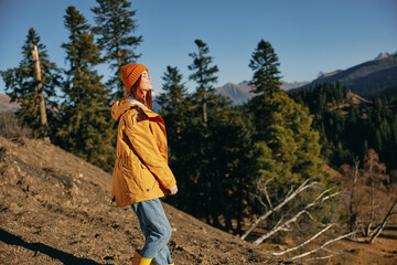 Naklejka premium Woman traveling to the mountains in the fall on a nature hike smile and happiness in a yellow cape with red hair full-length stands against the backdrop of trees and mountains