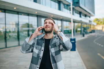 In the city, an anxious young bearded man is talking on the phone with a serious look. Sad Guy Engaging in Emotional Phone Communication in the City - obrazy, fototapety, plakaty