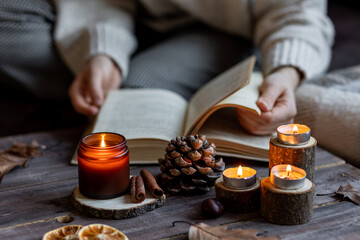 Cozy autumn or winter composition with aromatic candle, wool sweater. Aromatherapy, home atmosphere of cosiness and relax. Woman reading a book. Wooden background close up.
