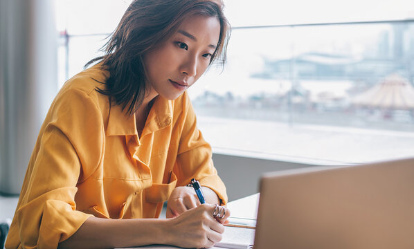Asian female writing and looking at screen display at workplace