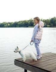 A seven-year-old girl with a small white dog stands on a bridge near the lake and looks at the water.