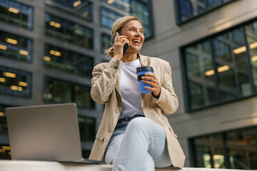 Inspired woman manager talking with client and work on laptop on modern office building background