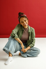 Charming stylish woman sitting on floor and looks camera with smile over red studio background