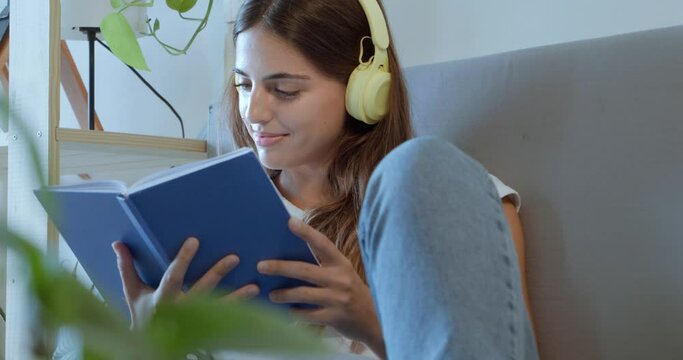 Young woman reading a book happy while listening to music with headphones lying on the sofa in an apartment