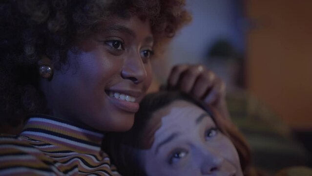 Young romantic couple girls lying at home watching TV. Afro woman affectionate caresses her Caucasian partner head as enjoy night of domestic life. Happy and loving LGBT relationships. Close up people