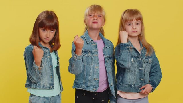 Aggressive angry teenage girls trying to fight at camera, shaking fist, boxing with expression, punishment, abuse, bullying. Little children sisters. Three siblings kids isolated on yellow background