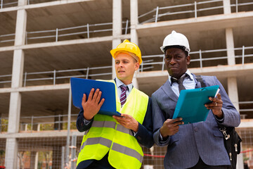 African-american foreman and Caucasian engineer in hardhats standing on construction site and talking about new building. One man using laptop, another one holding documents in hands.