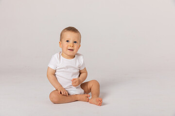 Little kid in white bodysuit, barefoot is smiling, sitting on floor against white studio background. Concept for articles about childhood or advertising for babies. Close up
