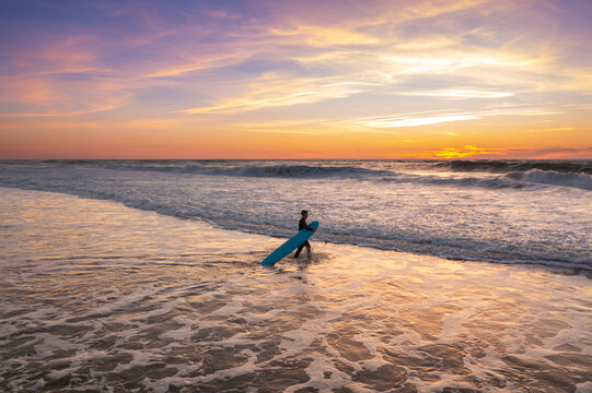 Surfer walking in ocean on sunset with surfboard. Person with surfboard stands in ocean water. Landscape with pink and violet clouds, ocean waves and sand beach. Surfer wathing the waves 