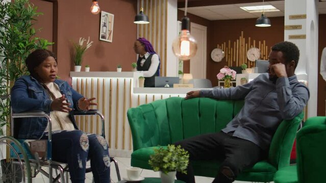 African american guest talking to man while she sits in wheelchair, entering disability friendly hotel lobby. Couple travelling abroad, reduced mobility tourism industry.