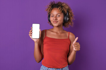 Young positive beautiful African American woman in headphones holds phone and shows thumb up listening to good music or audio books through mobile application stands in purple studio. White screen
