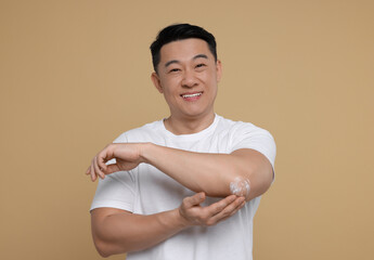 Handsome man applying body cream onto his elbow on light brown background