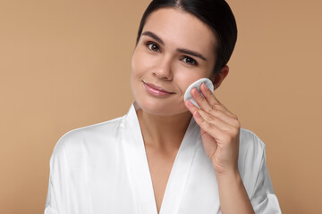 Young woman cleaning her face with cotton pad on beige background