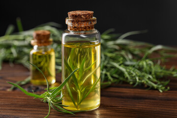 Bottles of essential oil and fresh tarragon leaves on wooden table, closeup. Space for text