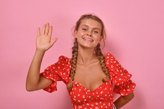 Young positive friendly Caucasian woman waves hand and says hello at meeting acquaintance or expresses hospitality to invite you to participate in promotion stands posing in pink studio. Hi, welcome