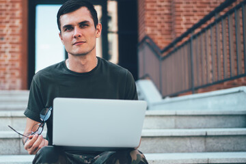 Calm cool man interacting with laptop while sitting on stairs on street