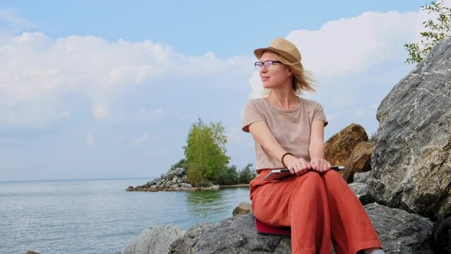 Blonde woman wearing hat sits on the rock near the sea using a laptop, working remotely. Happy female writer looking for inspiration in the seascape.