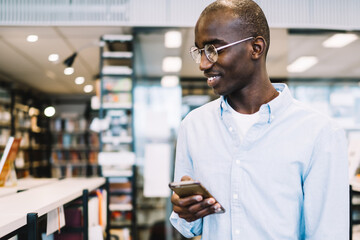 Black man walking along bookcases with smartphone and smiling