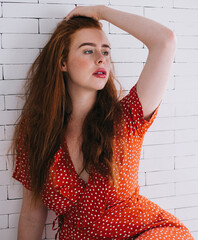 Confident woman with bright hair in red dress sitting near white brick wall