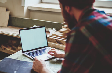 Faceless guy in casual shirt working with blank screen laptop at workshop