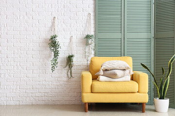 Yellow armchair with cushions and green folding screen near white brick wall