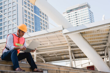 Construction engineer using laptop and sitting outdoor site building working blueprint project,...