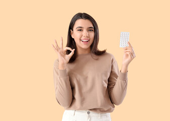Young woman with birth control pills showing OK on beige background