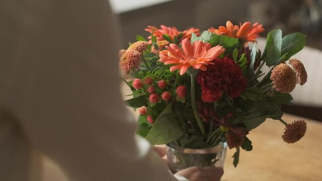 Putting vase with orange flowers on wooden table by female hands, slow motion