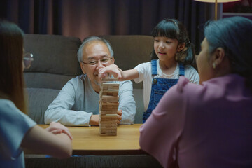 Happy grandparents Asian family enjoy playing toy block with little daughter and mother together at home night time, Smiling parent having fun play build constructor tower of wooden blocks, education - obrazy, fototapety, plakaty