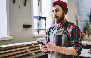 Calm adult woodworker pondering while using smartphone in modern workroom