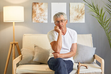 Bored sad upset grey-haired mature man wearing casual white T-shirt sitting on sofa at home...