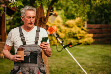 A mature male finishes trimming the lawn, peeling off his work attire, and enjoys a coffee to go while texting on the phone