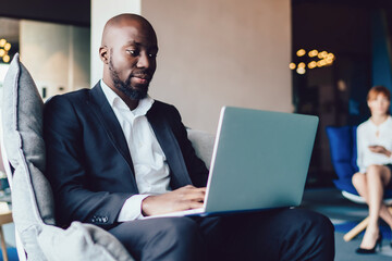 Concentrated black male executive in suit browsing laptop at lobby of business center