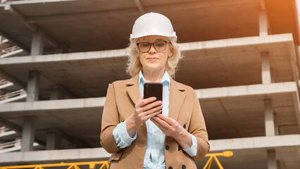 Blonde architect in protective helmet holds phone. Mature businesswoman reports about building...